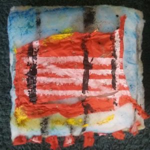 Image shows: square white cushion with 8 small red prayer flags sewn to bottom edge. A white gate symbol is stenciled onto a fragment of red silk fabric song with three vertical black lines and hints of blue and yellow paint.