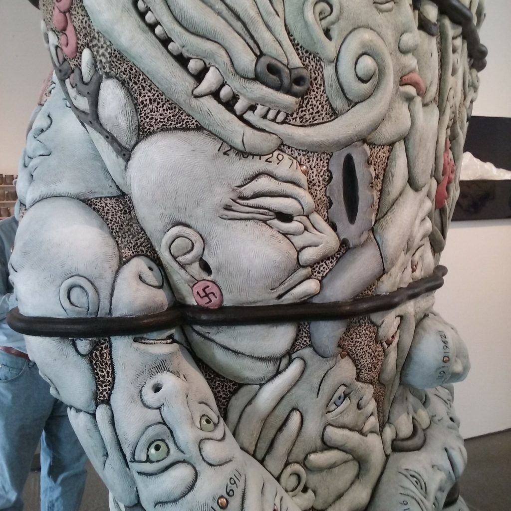 Matt clay faces with a folk art and political flavor carved into a tall column, pale colors and dark shadows in the revices