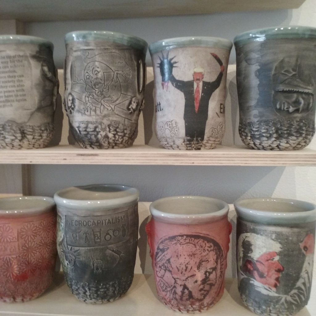 A detail of 8 cups on simple wooden shelves. One features an image of Trump holding a severed head of the Statue of Liberty, another a screaming Trump face. The other cups are subdued and have images of war impressed into them