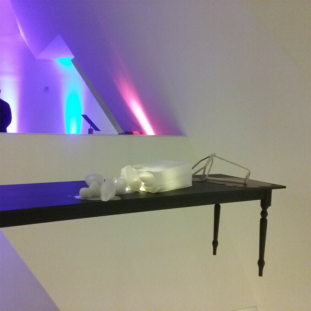 One side of The Enchanted, a sculpture featuring a long end of a low wooden table protrudes from a wall in mid air, with several white rubbery looking shapes placed on it, some are lit from within.