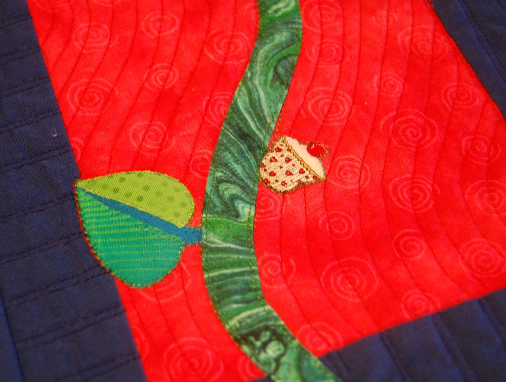 Detail of whimsical quilt featuring a cut-out of a cupcake added to green stem as if it were a fruit, with mottled green leaf to balance and a wavy quilting pattern over the whole design