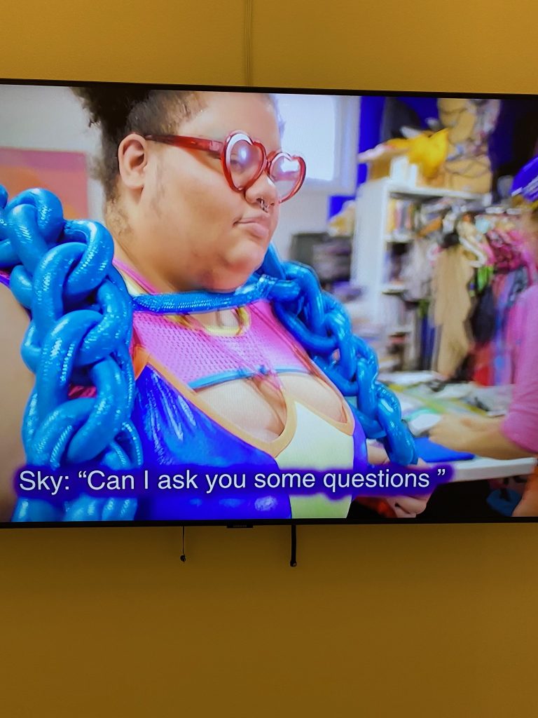 A detail of a video on a screen with text saying "Sky: "Can I sk you some questions" with a mixed race woman wearing red heart shaped glasses and an oversized metallic blue rope necklace with a body hugging lycra color blocked top with cut-outs. The background is a sewing workshop